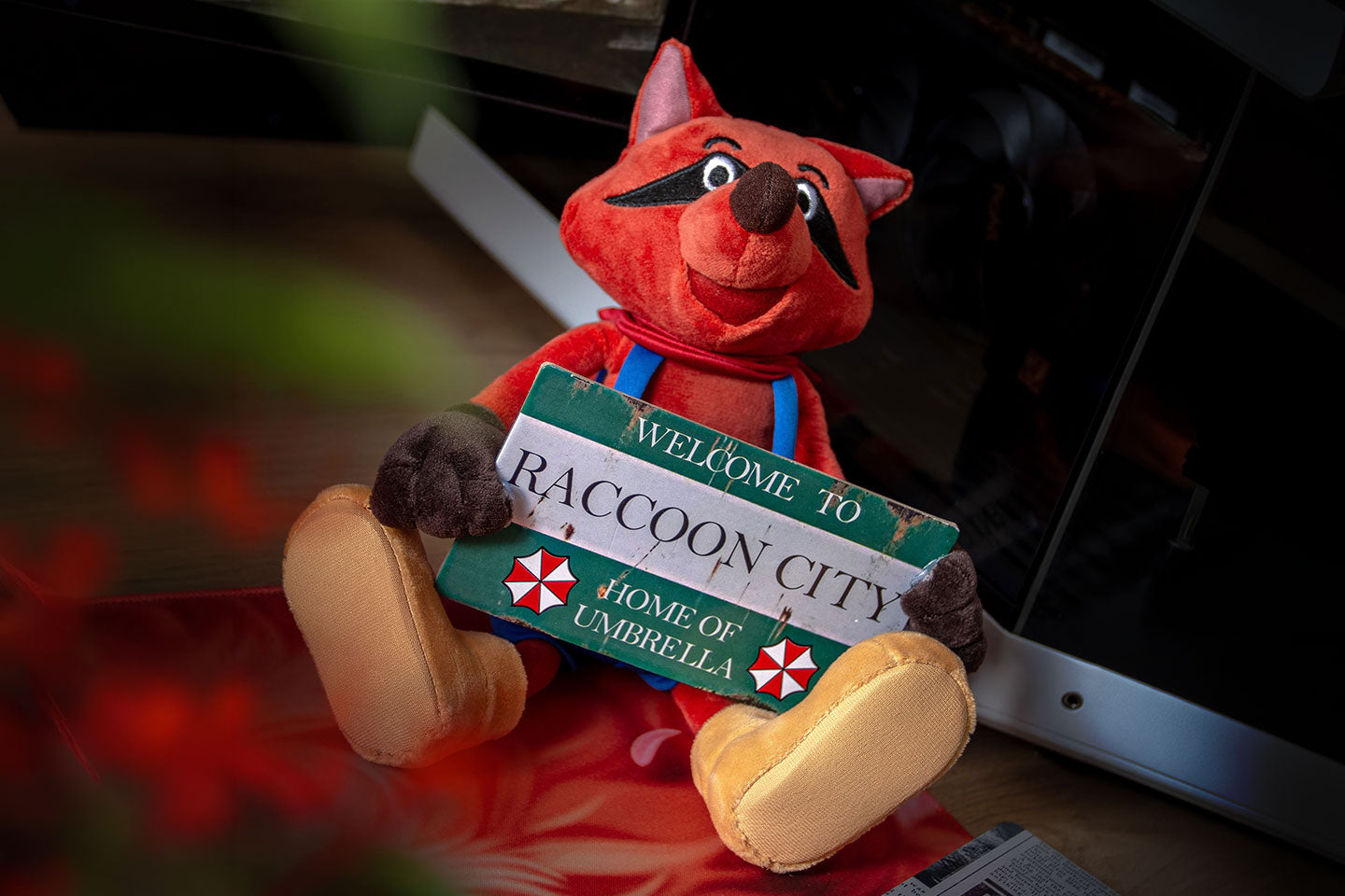 Resident Evil Mr. Raccoon Plüschfigur mit rotem Fell, blauer Latzhose und Welcome to Raccoon City Schild. Die Figur sitzt vor einem geöffneten Laptop in einer dunklen Gaming-Atmosphäre.